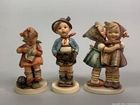 Front view of three Goebel Hummel figurines, showing a seated girl with bird, a standing boy, and two girls embracing.