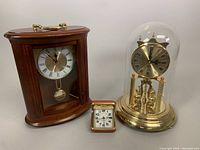 Three clocks including wooden case Linden mantel clock, brass bell jar Solar clock, and vintage red Westclox travel alarm clock arranged side by side.