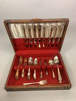 Flatware set neatly arranged in an open wooden box with red felt and white satin lining, showing variety of forks, spoons and butter knives.