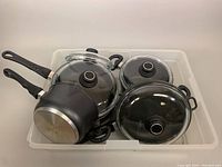 Overview of cookware in white plastic bin, including pots and pans with black handles and glass lids.