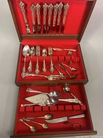 Top view inside wooden box showing multiple pieces of flatware arranged on red felt lining including cutters, spoons, forks, and specialty utensils.