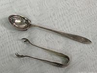Photo of teaspoon and sugar cube tongs laid flat on white textured fabric showing overall shape and condition