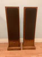 Front view of a pair of wooden cabinet floor-standing StudioLAB speakers with brown grille covers, standing side by side on a wood floor against a white wall.