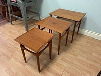 Three wood nesting tables in descending size order from left to right. Tables have light scratches and a warm wood finish. Hardwood floor and wall background.