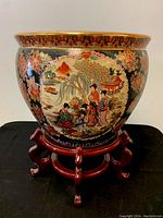 Front view of large hand-painted Satsuma planter pot on wooden stand, showing geisha scene and floral motifs.