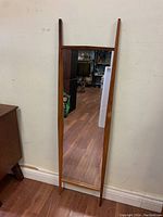 Full view of the Mid-Century Modern teak wall mirror leaning against a wall on wood flooring.