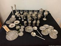 Wide view of full collection of pewter items on black cloth showing plates, mugs, goblets, ladles, candlesticks, coasters, and decorative dishes.