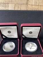 Two silver coins displayed in red presentation boxes, showing certificates inside the box lids.