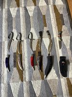 Six knives arranged horizontally on a patterned carpet. Five display cutters with curved blades and one hunting knife in sheath seen clearly.
