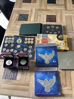 Photo showing seven sets of Canadian coins from 2000 including blue Celebrate the Year 2000 sets, black cases with silver coins, a green binder folder, a black coin portfolio with provincial shields, and a yellow box.