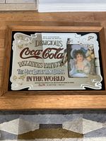 Front view of the vintage Coca-Cola mirror sign showing detailed Coca-Cola text and the image of a woman holding Coca-Cola bottles on the right side within a wood frame.