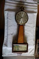 Front view of the vintage banjo clock showing brass eagle atop, clock face, birds eye maple panel, and painted house image at the bottom