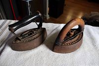 Photo of two antique sad irons side by side, showing both wood handles and cast iron bases with visible rust and wear.