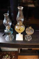 Three old oil lamps displayed on a dark surface showing their overall sizes and conditions.