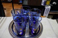 Five cobalt blue glass cups decorated with gold stars arranged on a round silver plated tray. The tray has a beaded edge.