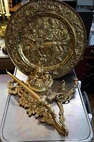 Large brass wall relief plate with an ornate border and raised scene of people, along with brass wall shelf and candle holder arranged on a metallic surface.