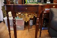 Front view showing the demi-lune shape, wood finish, drawer, and ornate gold trim underneath.