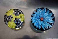 Top view of two glass paperweights, one with yellow and black flowers, the other with a blue flower and red center.