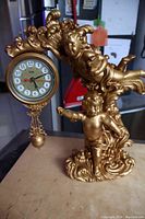 Side angle showing two gold cast resin putti figures and hanging clock face with pendulum ornament.