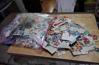 Large clear ziplock bag filled with loose postage stamps and an adjacent pile of stamped paper pieces including many different stamp designs.