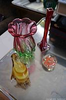 Four Mid-Century Modern art glass pieces displayed on a reflective surface including ruffled vase, tall red vase, penguin figurine, and clear paperweight with orange pattern