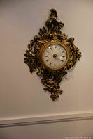 Full view of the antique French wall clock mounted on wall showing ornate gold-tone cast brass frame and porcelain dial with Roman numerals.