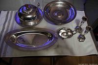 Photo of the entire silver plate lot including covered butter dish, serving bowl, oval tray, candlesticks, and ladle bowl on a cloth surface.