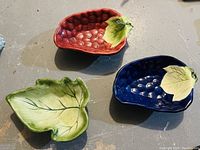 Three ceramic decorative bowls shaped as fruit and leaf, including a red strawberry bowl, blue grape bowl, and green leaf bowl placed on a floor.