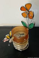 Glass round container with metal base, orange flower and green leaves attached by metal wire, and butterfly decor attached to the side