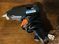 Photo showing a compact black electric glue gun with an orange trigger, attached power cord wrapped around the handle, and a label on the side reading "Glue Gun XG-11".