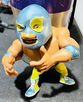 Close-up photo of a PVC wrestling toy figure wearing a blue mask and yellow-blue wrestling outfit standing on a black surface.
