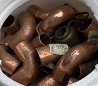 Close-up photo of various copper pipe fittings inside a white bucket, showing 90-degree elbows and straight connectors.
