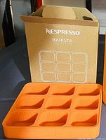 Orange silicone storage tray with 12 compartments for Nespresso pods next to original Nespresso Barista Inspirations box.