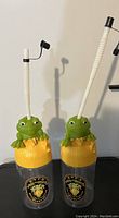 Pair of clear plastic tumblers with yellow lids shaped like grassy stumps and green frogs on top, with white flexible straws attached.