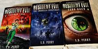 Three Resident Evil paperback books by S.D. Perry displayed flat on a surface: 'Caliban Cove', 'The Umbrella Conspiracy 1', 'Zero Hour'. Covers show respective artwork and author name clearly.