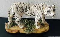 White tiger figurine shown from the side, displaying detailed black stripes, blue eyes, and a textured grass and earth colored base.