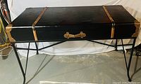 Photo of a black suitcase-style table with leather-like straps and brass hardware on black metal legs in a concrete floor setting.