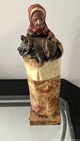 Photo of vintage paper mache figurine showing an elderly woman with a headscarf holding a pig, sitting on a marbled pedestal.