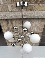 Front view of 8 bulb space age chandelier with chrome finish and white frosted globe bulbs against brick wall background.