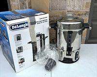 Full view of the DeLonghi coffee urn next to its original box on a table outside, showing the urn, box, plastic power cord wrapper, and tap dispenser clearly.