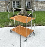 Full view of folding rolling bar cart showing wood-grain trays and chrome frame on caster wheels.