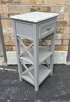 Side and angled view of small gray wooden cabinet with marble top, drawer, and shelves against brick wall.