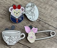Four Disney collector enamel pins laid out on wooden surface: Hidden Mickey Tea Cup pin, Piglet pin with pink ears, white bear in sailor outfit with red bow pin, and silver ballet dancer pin.