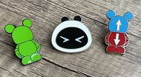 Photo showing three Disney enamel collector pins: green Mickey Mouse figure from behind, stylized white and black face, and red/blue Mickey Mouse figure with arrows on ears and body.
