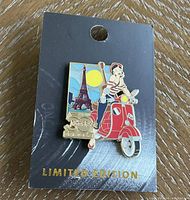 Close-up of a limited edition Hard Rock 50th Anniversary Girl Series 21 collector pin featuring a girl on a red Vespa against a Paris themed backdrop, mounted on a black card with gold text.