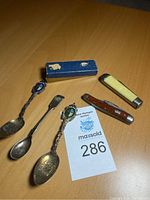 Photo showing three antique spoons, two marked sterling and one with hallmarks, along with two pocket knives and a small blue box.