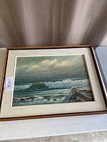 Full view of the framed original oil painting showing ocean waves crashing against rocks and a cloudy sky.