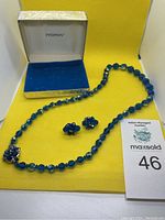 Blue beaded necklace laid out with matching earrings on yellow background with open Peoples box.