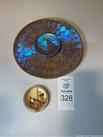 Two round plates featuring Blue Morpho butterfly wing patterns with central magnifying glass, one with brighter blue hues and brass rim. Also a round clear resin paperweight with a preserved beetle and plant inside.