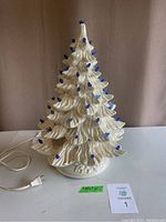 Full view of the vintage white ceramic Christmas tree lit with blue lights attached through the branches. The tree is plugged in with a visible white power cord.